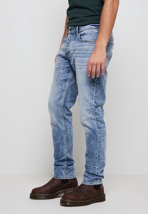 Homme portant un jean bleu clair délavé et des bottes en cuir marron foncé, debout devant un fond uni.