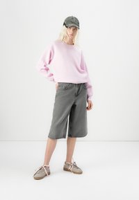 Felpa rosa, pantaloni bermuda in denim grigio e sneakers grigie con lacci rosa. Il cappellino è di colore verde scuro con visiera curva.