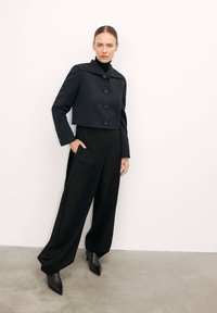 Fekete, rövidített kabát széles gallérral és gombokkal, magas derekú fekete nadrággal párosítva. A modell hegyes orrú fekete bokacsizmát visel.