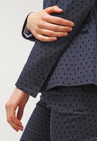 Costume bleu marine à pois avec une coupe ajustée ; doté de boutons sur la manche et d'une texture lisse. Comprend des détails de pois foncés sur le tissu.