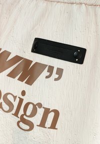 Tejido de textura crema con un prominente diseño gráfico marrón y una pequeña etiqueta de cuero negro con marca grabada.