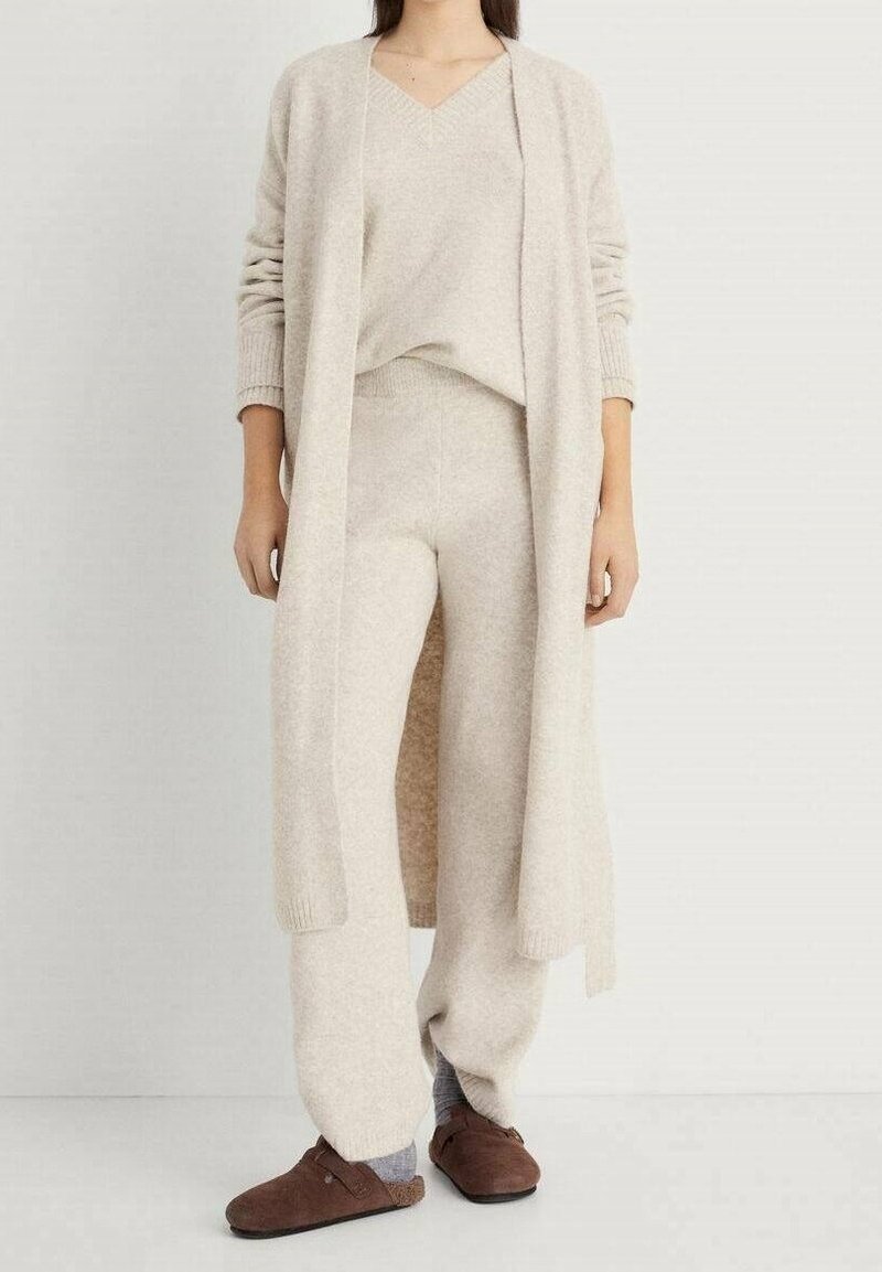 Femme portant un haut en maille beige à col en V, un pantalon en maille assorti, un long cardigan ouvert, des chaussettes grises et des sabots marron sans lacets, debout sur un sol blanc.