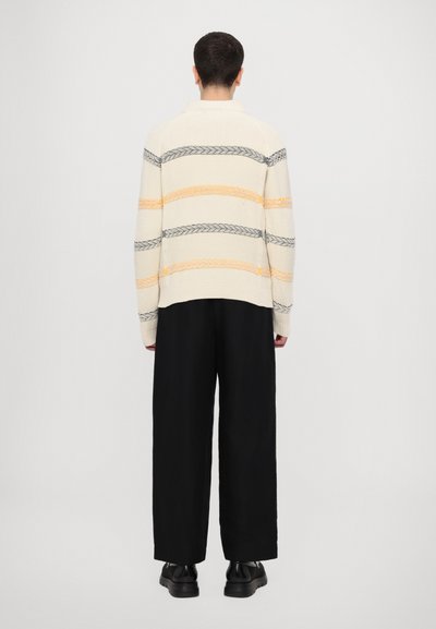 Pull en tricot crème avec des rayures horizontales gris et orange, doté un col montant et une coupe décontractée. Associé à un pantalon noir à jambes larges.