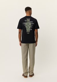 Μαύρο κοντομάνικο μπλουζάκι με πράσινο σχέδιο ενός περίτεχνου βάζου και κείμενο στην πλάτη. Συνδυάζεται με ανοιχτόχρωμο παντελόνι.