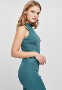 Ung kvinde med lyst r�ødt hår iført en teal-ribbet ærmeløs top og matchende bukser, poserer sidelæns mod en lys, ensfarvet baggrund.