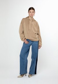 Sudadera beige con capucha, bolsillo frontal y logotipo; combinada con jeans de mezclilla azul de pierna ancha con rayas laterales y zapatos metálicos de punta.