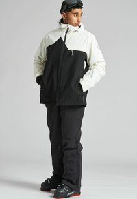 Chaqueta de nieve en blanco y negro con un diseño geométrico, combinada con pantalones de nieve negros y botas negras con acentos rojos.