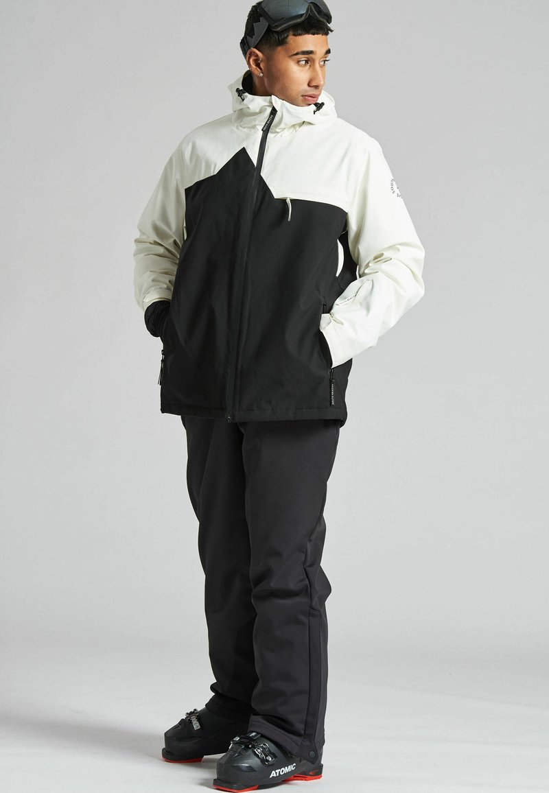 Chaqueta de nieve en blanco y negro con un diseño geométrico, combinada con pantalones de nieve negros y botas negras con acentos rojos.