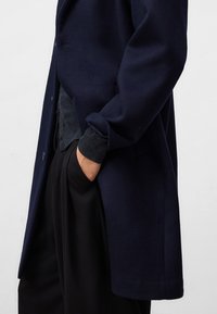 Marineblauer Wollübermantel mit einem maßgeschneiderten Design, ausgestattet mit einer einzelnen Knopfschließe und umgeschlagenen Bündchen. Tragen über einem dunkel strukturierten Hemd und lockeren schwarzen Hosen.