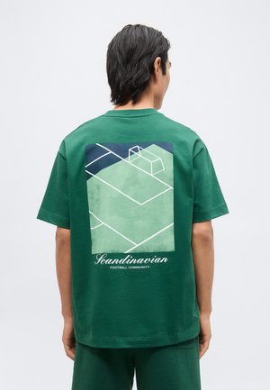 Homme de dos portant un t-shirt vert avec un graphisme minimaliste de terrain de football et le texte "Scandinavian Football Community".