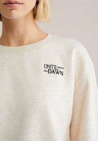 Svetlo bež jopica z teksturirano obdelavo. Na levi strani prsnega koša je črni vezeni napis "UNTIL DAWN". Dizajn z okroglim vratom.