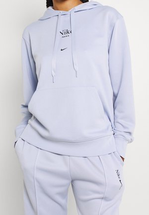 Person iført lys lilla Nike-hoodie og matchende joggers med frontlomme og Nike-logoer på bryst og lår.
