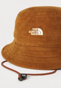 Brun corduroy bøttehat med hvidt logo. Har struktureret ribbet stof, en bred skygge og en justerbar brun snor.