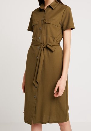 Woman wearing a knee-length olive green shirt dress with short sleeves, front buttons, chest pockets, and a tied waist belt.