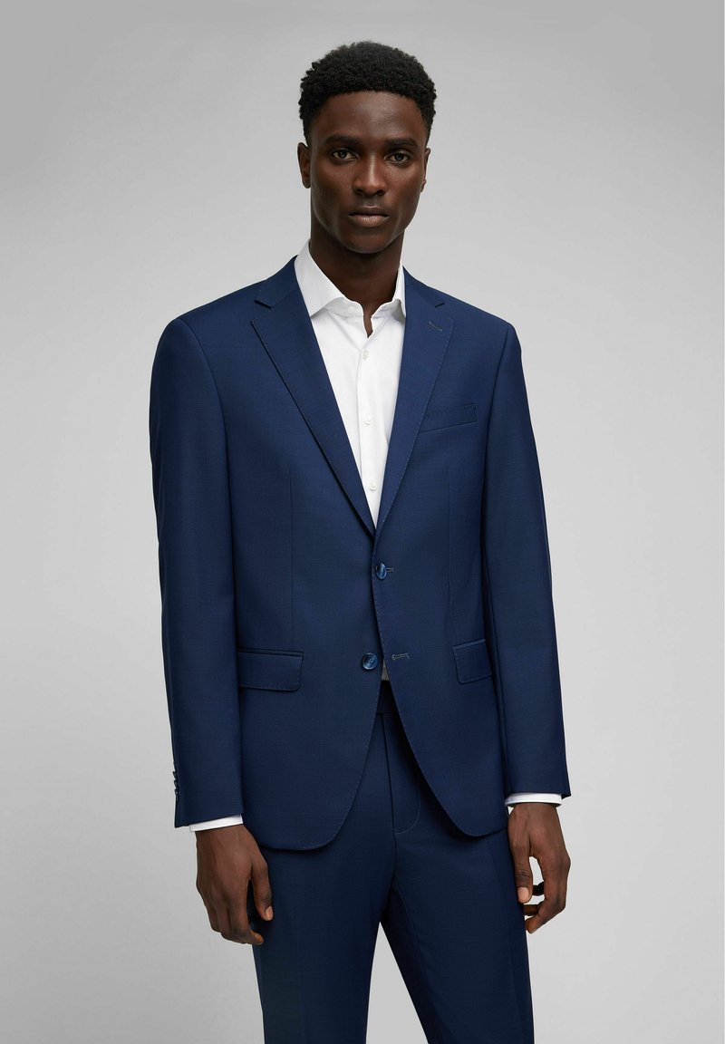 Abito blu navy con chiusura a due bottoni, rever a punta e tasche frontali. Abbinato a una camicia bianca con colletto in tessuto testurizzato.