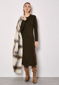 Grünes, gestricktes Langarmkleid mit taillierter Silhouette, kombiniert mit einem creme- und braunkarierten Schal. Knielange, tanfarbene Stiefel mit glatter Textur und stilvollem Design.