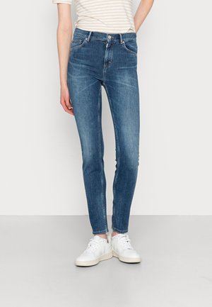 Osoba w niebieskich dopasowanych jeansach, białych tenisówkach i jasnej, prążkowanej bluzce z krótkim rękawem, stojąca na tle jednolitego jasnego tła.