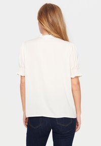 Maglietta bianca a maniche corte con polsini arricciati, vestibilità comoda e scollatura rotonda. Il modello indossa jeans blu scuro, mostrando un look casual.