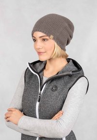 Grijze gebreide beanie met een losse pasvorm en een ribbeltextuur. Heeft een klein logolabel. Gecombineerd met een grijze sweatervest met zwarte accenten.