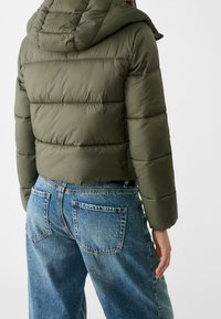 Olivgrön vadderad jacka med huva och sydda paneler, kombinerad med ljusblå vida jeans med bakfickor.