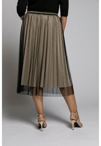 Femme portant une jupe beige avec un superposition en maille à pois noirs et des sandales à talons dorés, se tenant contre un fond gris uni.