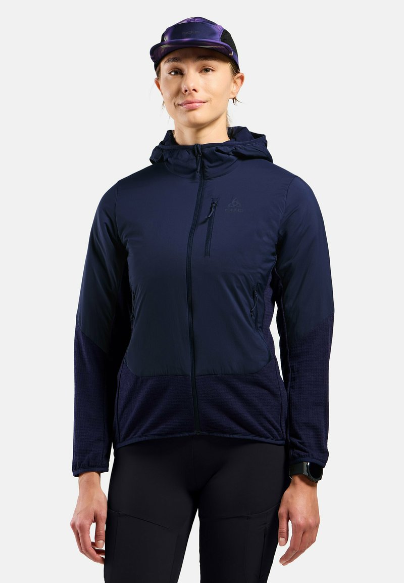 Marineblauwe hoodie met gestructureerde mouwen, ritszakken en een getailleerd ontwerp. Draagt een zwart horloge en een paarse pet.