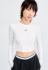 Langermet hvit crop topp laget av mykt stoff, med en svart Nike-logo på brystet, paired med svarte joggebukser med merket i livet.