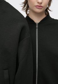 Zwarte bomberjas met een zilveren rits, geribde kraag en gestroomlijnd ontwerp. Zacht materiaal met een strakke, minimalistische stijl.