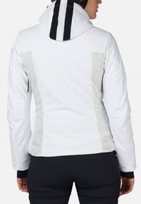 Giacca impermeabile bianca con design aderente, dotata di cappuccio a strisce nere, pannelli laterali trasparenti e tasche con zip.