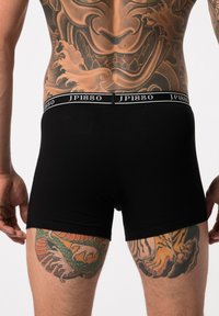 Caleçons boxers noirs avec une taille douce arborant le logo "JP1880", texture lisse et coupe ajustée, accompagnés d'un art corporel vibrant sur les cuisses.