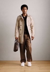 Uomo che indossa un trench beige, pantaloni marroni, sneakers bianche, mentre porta una valigetta marrone scuro, in piedi su un pavimento di legno contro un muro semplice.