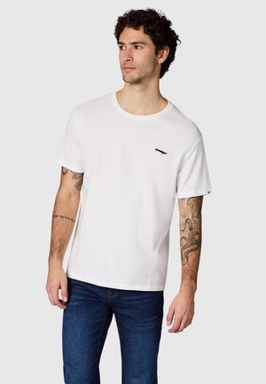 Jonge man met krullend haar die een effen wit T-shirt met een klein zwart logo en een blauwe spijkerbroek draagt, die zijwaarts kijkt.