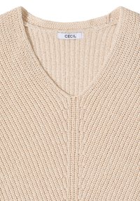 Pull tricoté couleur crème avec un col en V et une texture côtelée, comportant une étiquette blanche avec la marque "CECIL" à l'intérieur du col.
