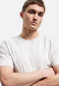 Hvid bomulds-T-shirt med rund halsudskæring og korte ærmer. "DUBLIN" er trykt i lys grå på forsiden. Glat tekstur.