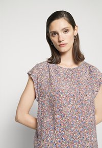 Mulher com cabelo castanho escuro, usando brincos de argola prateados e uma blusa de mangas curtas com estampado floral, com flores azuis e beges sobre um fundo vermelho.