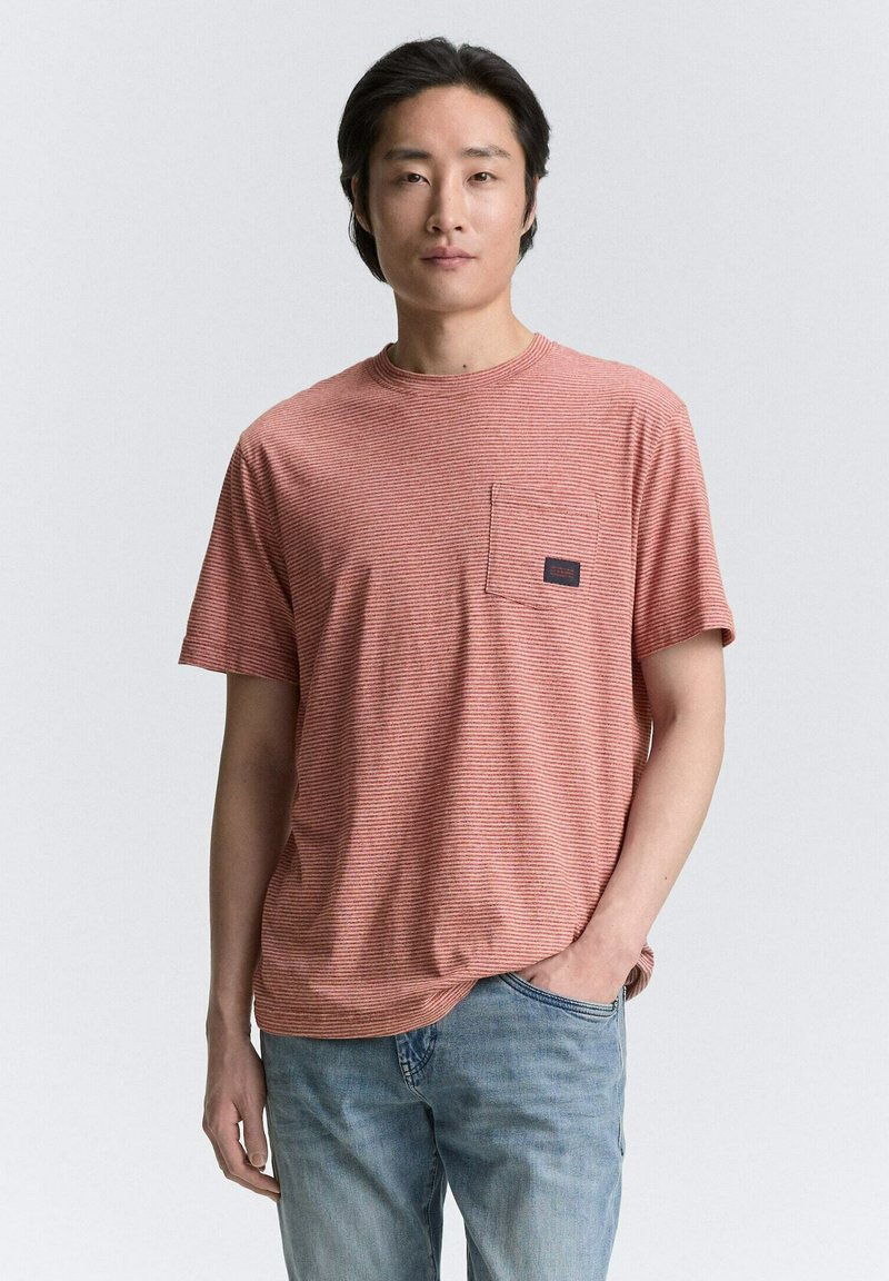 Gestreept oranje t-shirt met een ronde hals, korte mouwen en een linker borstzak met een donker label. Draag het met lichtblauwe spijkerbroeken.