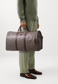 La borsa duffel tessuta presenta un motivo geometrico in tonalità di viola e marrone, con manici in pelle marrone scuro e dettagli in metallo dorato.
