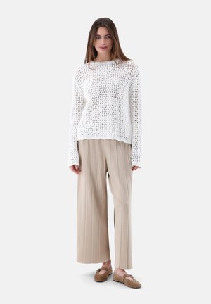 Femme portant un pull en maille blanche ajourée, un pantalon beige à larges jambes plissé et des ballerines marron clair, debout devant un fond clair uni.