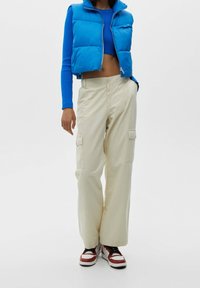 Chaleco azul tipo puffer con cierre de cremallera, top corto azul de canalé, pantalones cargo beige con bolsillos y zapatillas rojas y blancas.