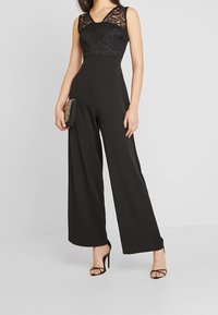 Jumpsuit de encaje negro con diseño de pierna ancha, que presenta un escote en v profundo y una tela suave. Un bolso minimalista y unos tacones con tiras completan el look.
