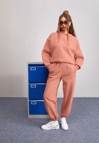 Conjunto de sudadera y pantalones de chándal en color rosa empolvado, fabricado en un tejido suave, con puños elásticos y cintura con cordón. Zapatillas blancas con detalles azules.