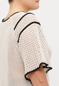 Knit top featuring an open weave in cream, with black trim along the neckline and sleeves, and scalloped edges. Textured, lightweight fabric.