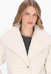 Beige steppelt dzseki nagy, hajtókás gallérral és egygombos záródással. Simább textúrával és egyszerű, modern designnal rendelkezik.