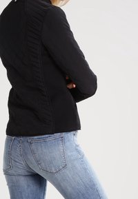 Svart vadderad jacka med en figursydd design, upprätt krage och texturerade sidodetaljer, i kombination med ljusblå denimjeans.