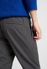 Szare, dopasowane spodnie z subtelną fakturą, z tylną kieszenią i wąskim krojem. Noszone z niebieskim swetrem dzianinowym.