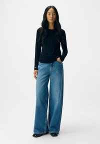 Sort langærmet top parret med brede blå denim jeans, der har høj talje og rene sømme; modellen står med den ene hånd i lommen.