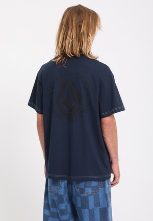 Personne aux cheveux mi-longs portant un T-shirt bleu foncé avec un logo circulaire délavé dans le dos et un short en jean patchwork bleu.