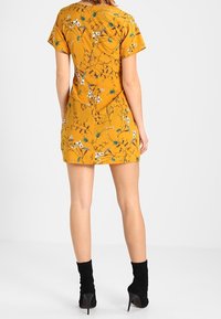 Sinappikeltainen mekko, jossa on lyhyet hihat, valkoisen ja vihreän kukkakuosi, vartalonmyötäinen siluetti ja vetoketjutakakappaleessa. Mustat nilkkurit.