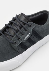 Tamsiai pilki sportbačiai su zomšos ir odos akcentais, su raišteliais, trimis juodomis juostelėmis ir balta gumine sola. Adidas logotipas matomas.
