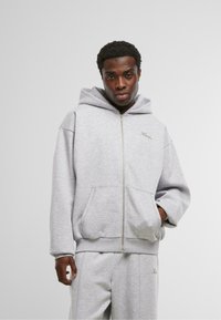 Felpa grigia con zip realizzata in materiale morbido, dotata di tasca frontale, polsini a costine e cappuccio, con un piccolo logo sul petto.